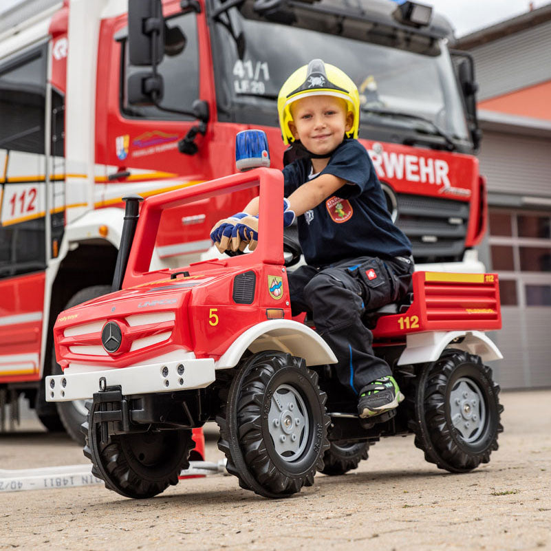 Child sits on Rolly Unimog Fire Truck in front of real fire truck.