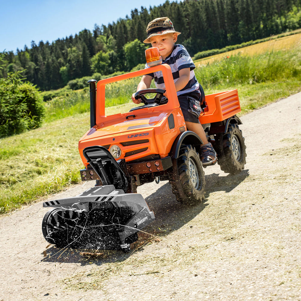Child driving an orange Unimog Service Truck toy with front loader on dirt path outdoors