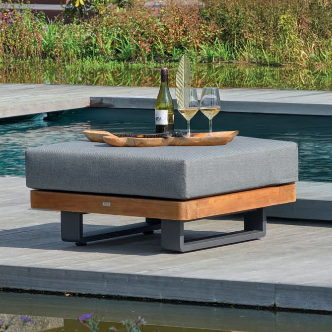 Fitz Roy Teak Ottoman with lava aluminum frame and charcoal cushion by a pool with drinks on top.