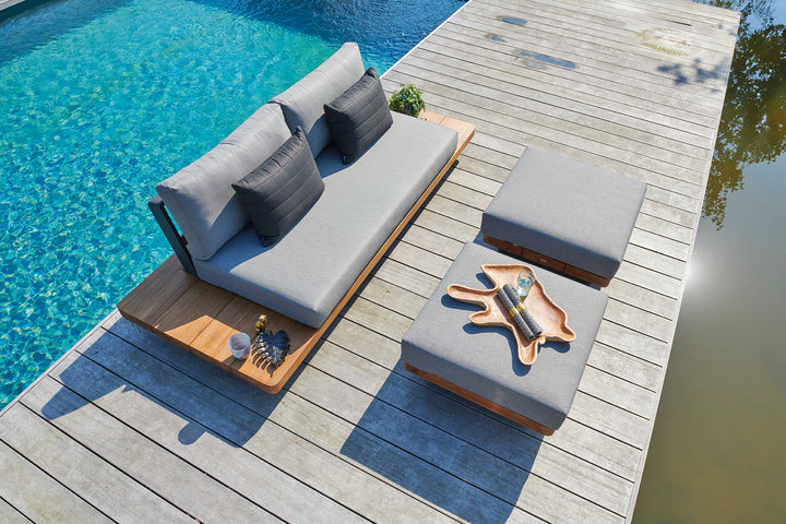 Fitz Roy Teak Ottoman with plush charcoal cushion and teak wood frame, set poolside on a wooden deck.