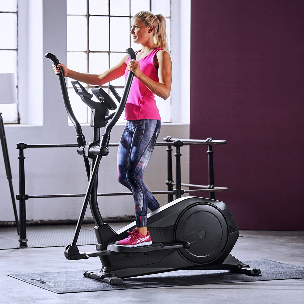 Woman exercising on the KETTLER Rivo 4 Elliptical Trainer with backlit display in a bright workout room.