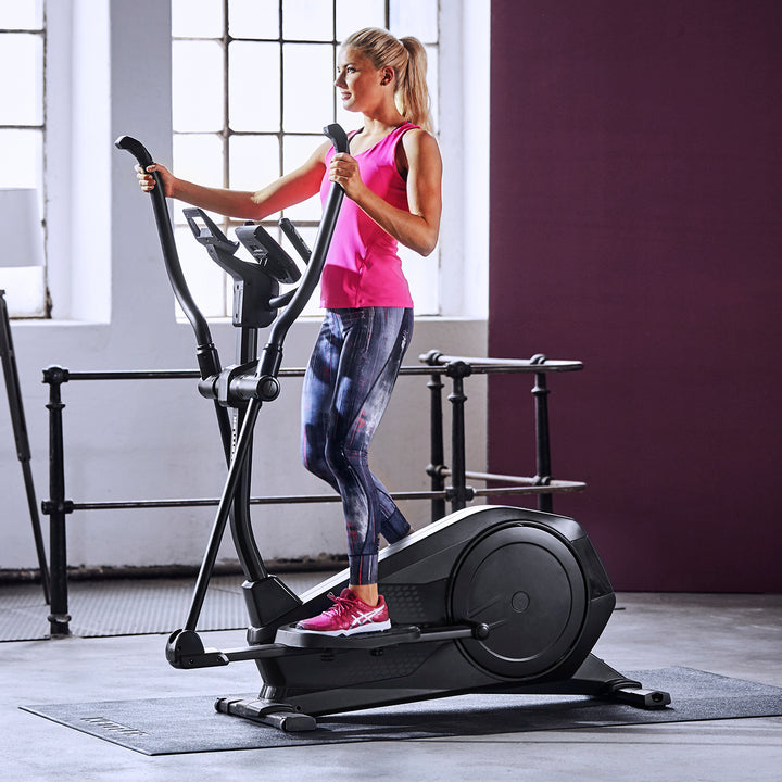 Woman exercising on the KETTLER Rivo 4 Elliptical Trainer with backlit display in a bright workout room.