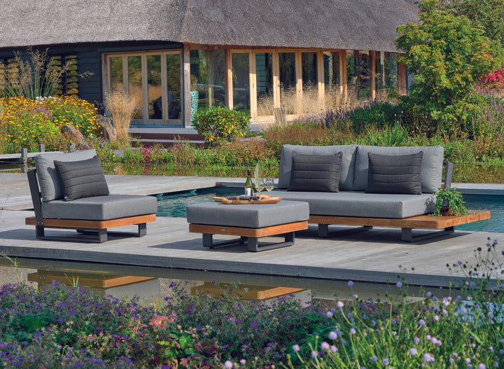 Fitz Roy Teak Ottoman with plush charcoal cushion and teak accents on durable lava aluminum frame by the pool.