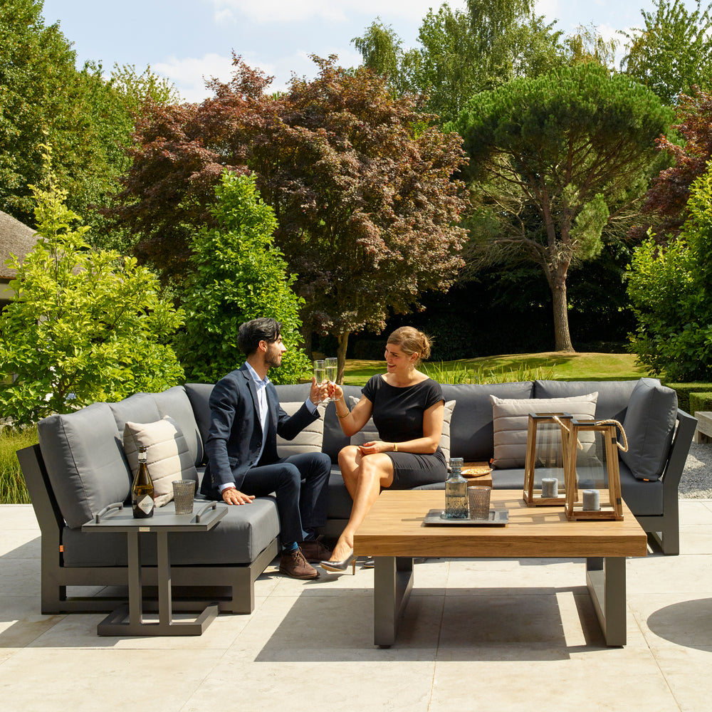 Nevada Small Corner Conversation Set with grey cushions and teak coffee table on outdoor patio with two people toasting.