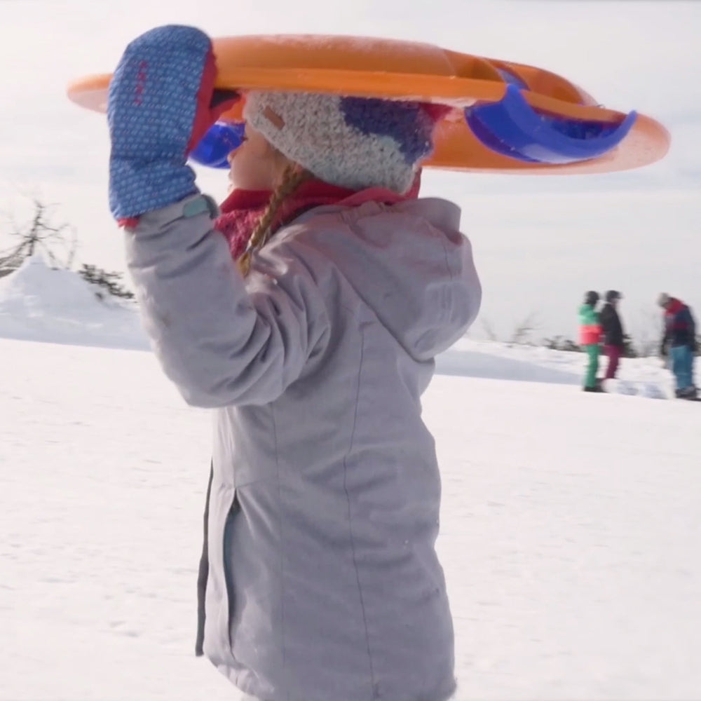 Child carrying orange Fun UFO Sled in snowy landscape wearing winter clothes and mittens.