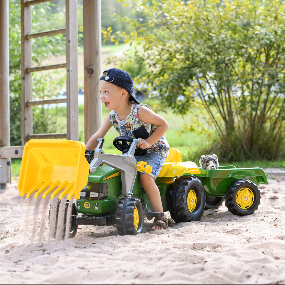 Child playing outdoors on a John Deere Pedal Tractor With Loader in a sandbox with a green trailer attached.