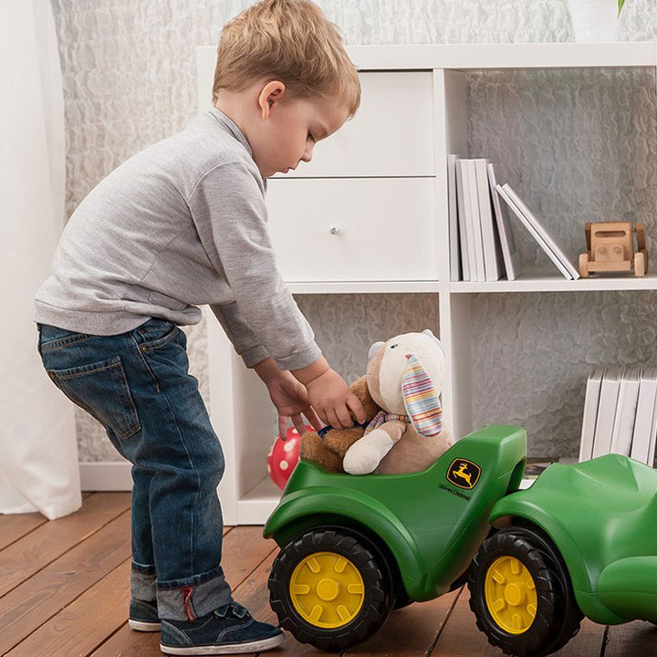 Child placing toys in green John Deere Minitrac Trailer attached to ride-on toy in a modern playroom.