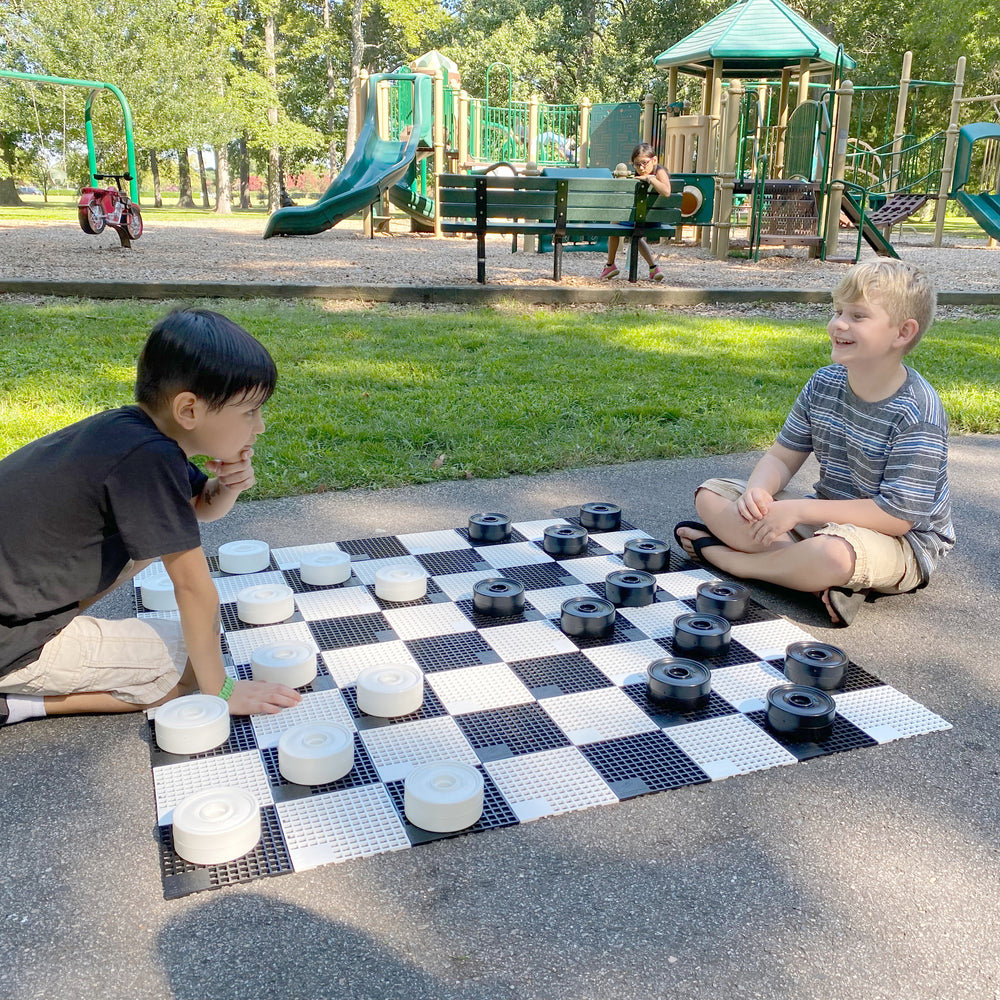 Children playing Mini-Giant Checker Pieces outdoors on a large black and white checkers board in a park setting.