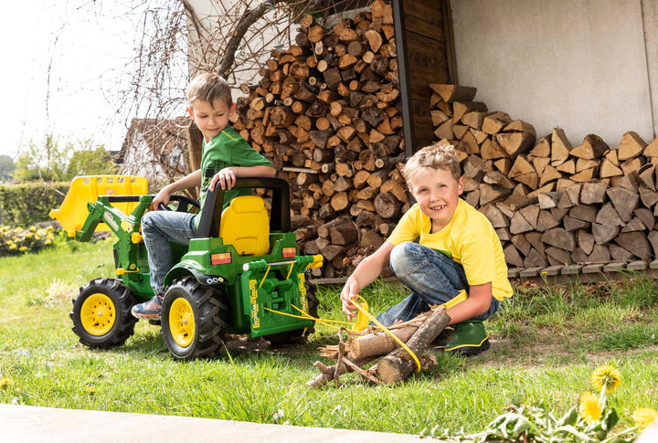 Two children playing with a toy tractor in front of stacked firewood.