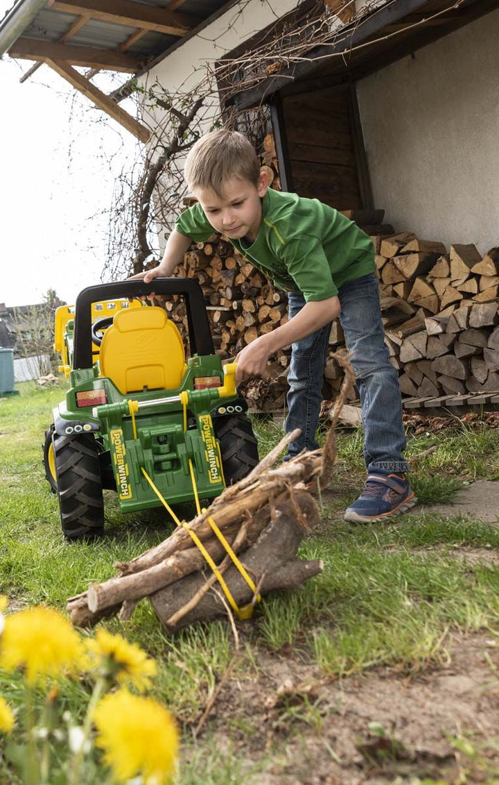 Child playing with a toy tractor pulling logs in front of a house with stacked firewood.