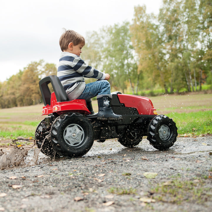 Child riding a red Case IH Tractor With Trailer splashing through a puddle outdoors on a dirt path.