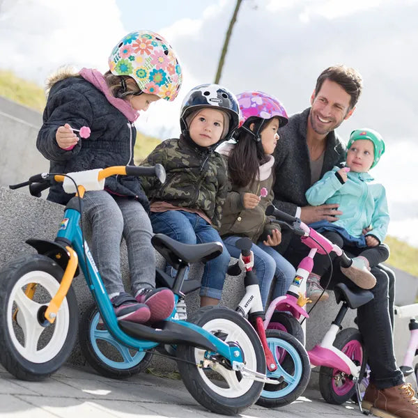 Children wearing helmets sitting on Layana Balance Bikes with an adult supervising outdoors.