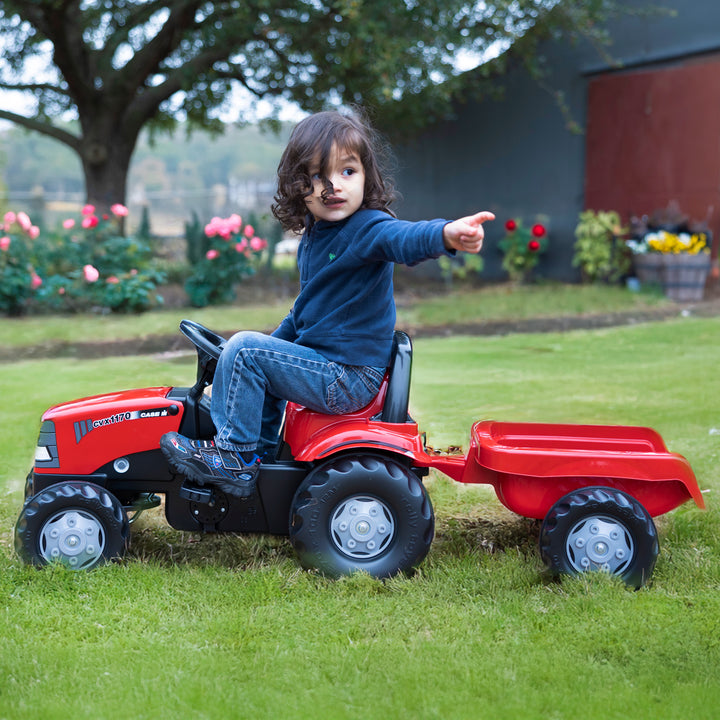 Child riding red Case IH Tractor With Trailer toy on grass in garden with trees and flowers in background