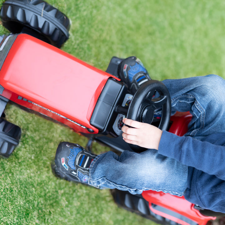 Child riding a red Case IH Tractor With Trailer toy on grass, featuring steering wheel and large replica tires.
