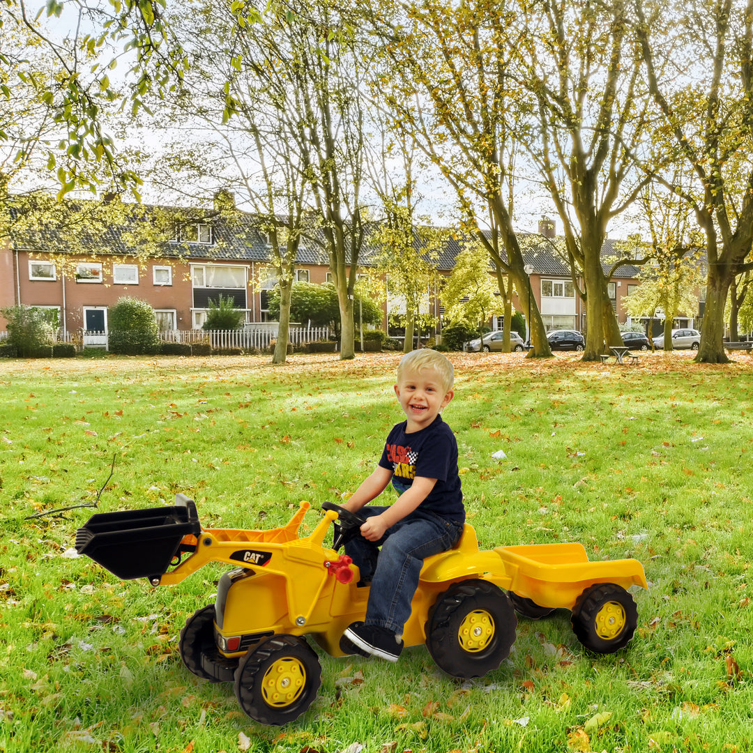 Child riding the yellow CAT Kid Tractor With Trailer pedal tractor with front loader and detachable trailer on green grass.