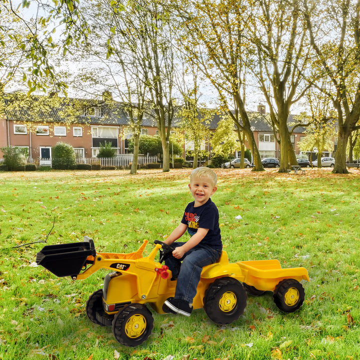 Child riding the yellow CAT Kid Tractor With Trailer pedal tractor with front loader and detachable trailer on green grass.