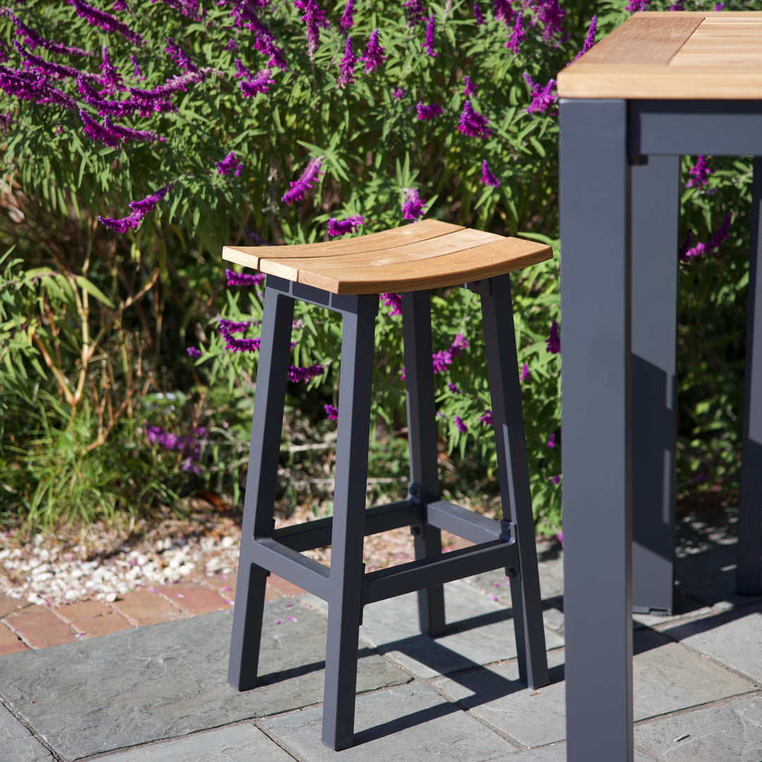 Elba Aluminum Bar Stool With Teak Accents featuring a charcoal frame and contoured teak saddle seat in an outdoor garden setting.