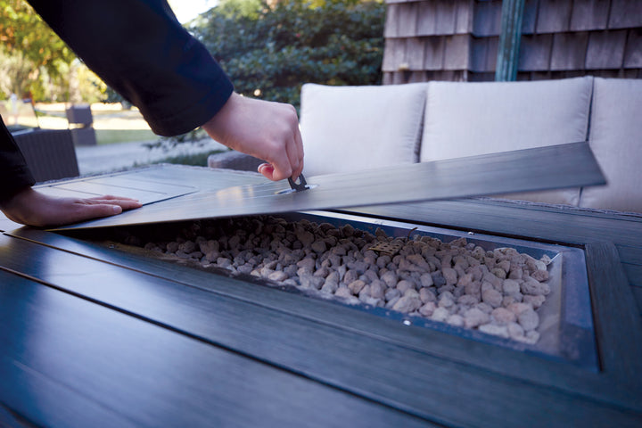 Person lifting lid on Palma Fire Table showing stone-filled firepit in outdoor setting.