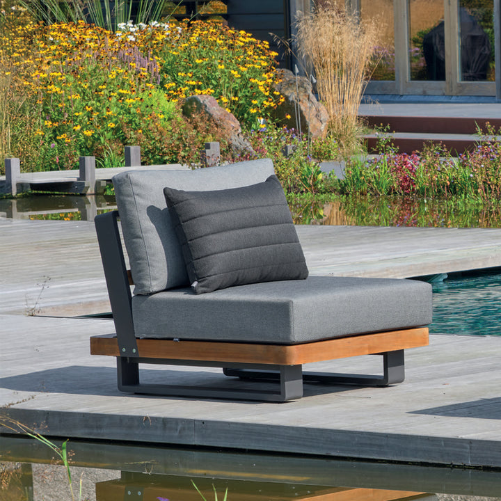 Fitz Roy Teak Armless Chair with teak frame, lava aluminum base, and charcoal cushions by a poolside garden.