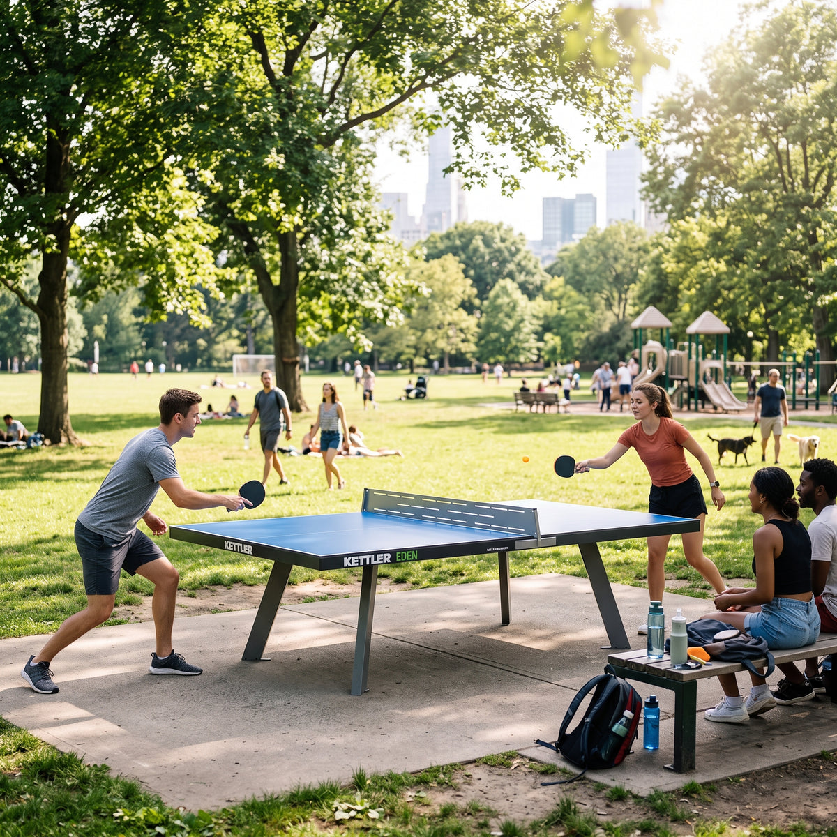 People play table tennis at an outdoor park table while spectators watch, with trees, a playground, and city buildings in the background
