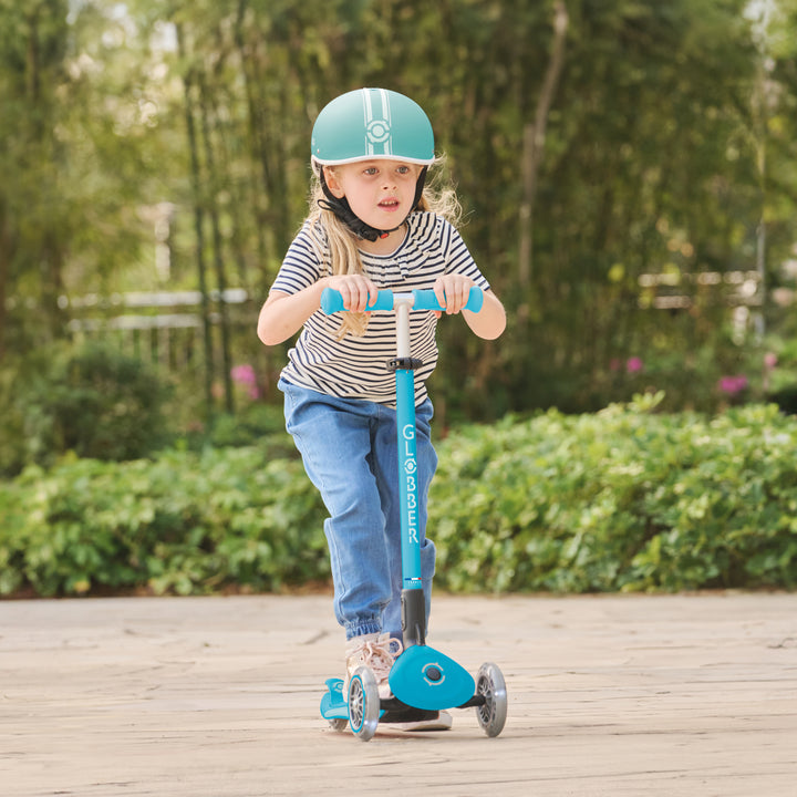 Child riding Aqua Elite Scooter with LED front wheels in park wearing helmet and striped shirt on paved path