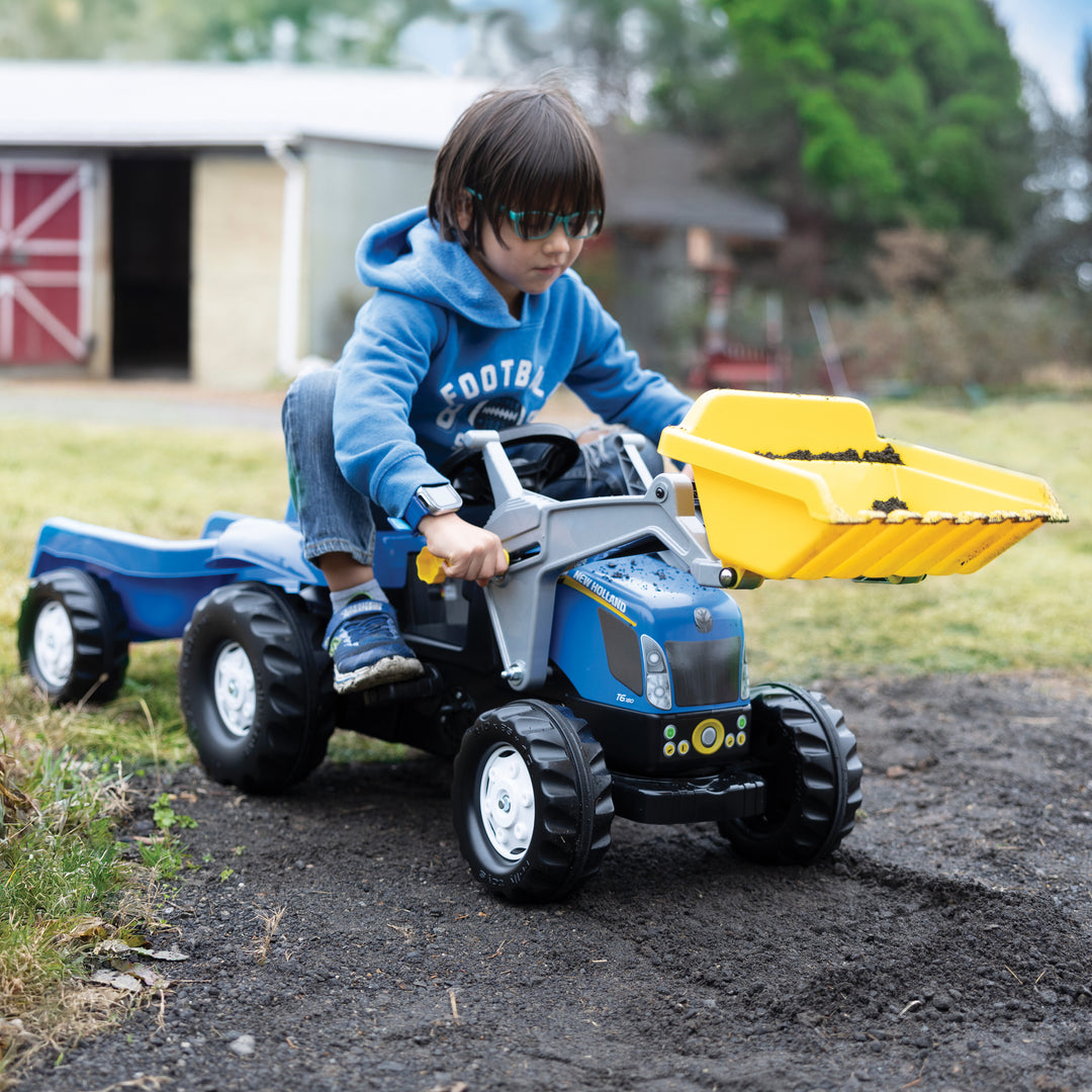 Child riding New Holland Front Load Kid Tractor w/Trailer with yellow front loader on dirt path outdoors.