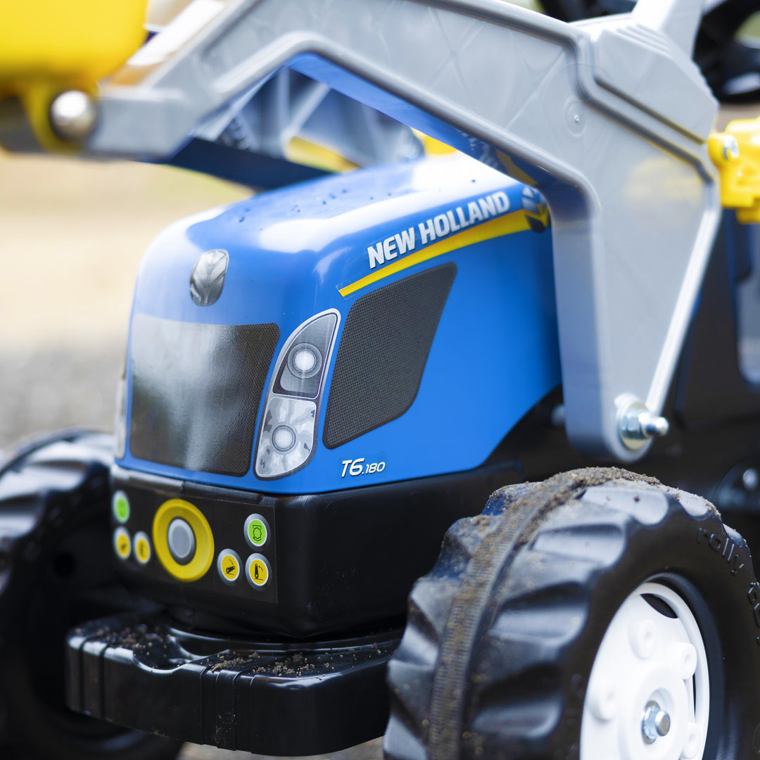 New Holland Front Load Kid Tractor w/Trailer close-up on blue hood and large black rubberized tires outdoors.