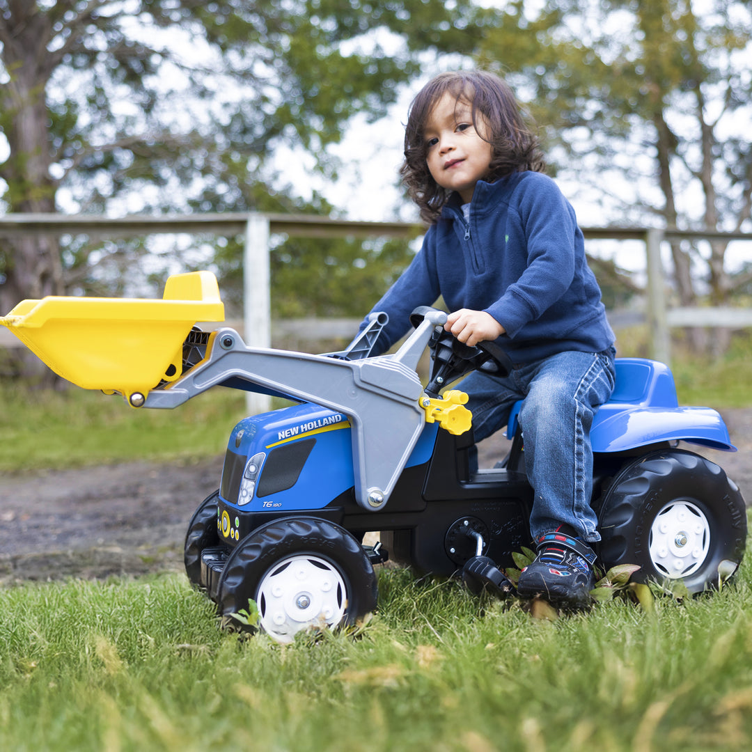 Child riding the New Holland Front Load Kid Tractor w/Trailer pedal tractor with yellow front loader outdoors on grass.