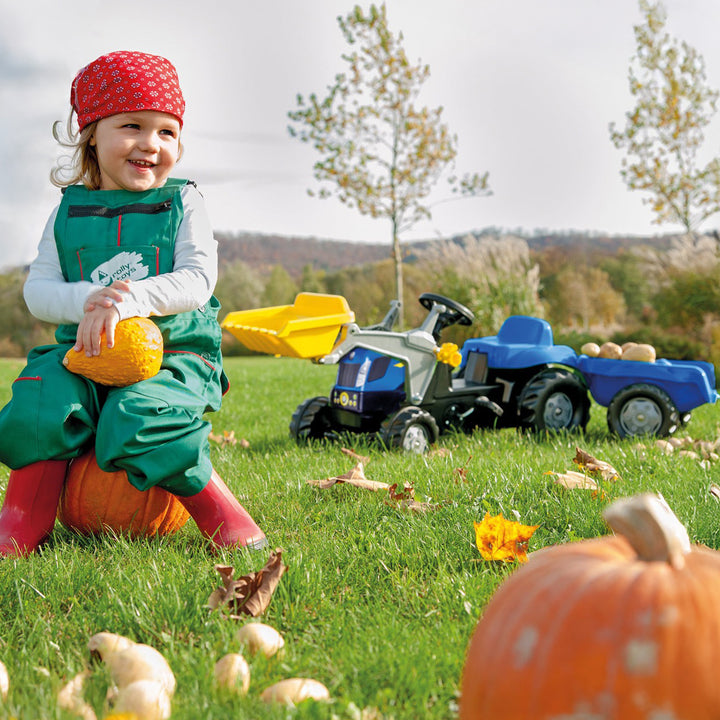 Child playing outdoors with New Holland Front Load Kid Tractor w/Trailer surrounded by pumpkins on grass.