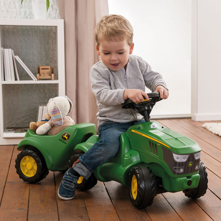 Child riding John Deere Minitrac Trailer attached to a green ride-on tractor indoors on wooden floor.