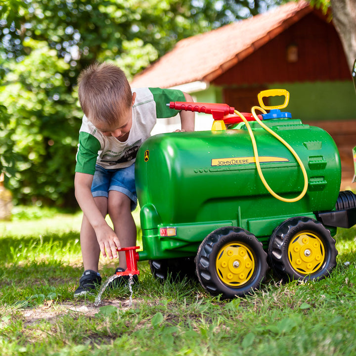 Child playing outdoors with John Deere Water Tanker With Pump & Spray Gun on grass near a wooden house