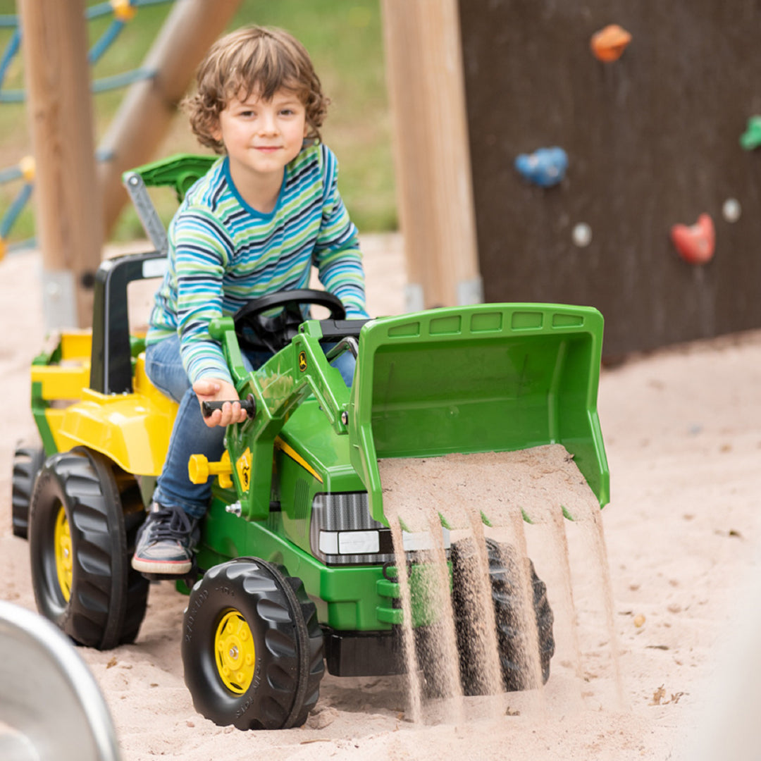 Child playing with John Deere Pedal Loader With Backhoe outdoor toy tractor in sandbox with sand pouring from loader bucket.