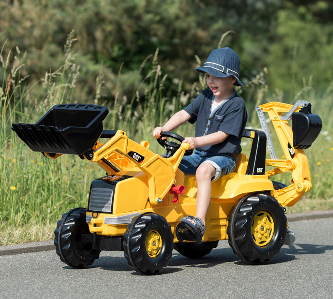 Child driving yellow CAT Front Loader With Backhoe Pedal Tractor outdoors on paved path with grass background