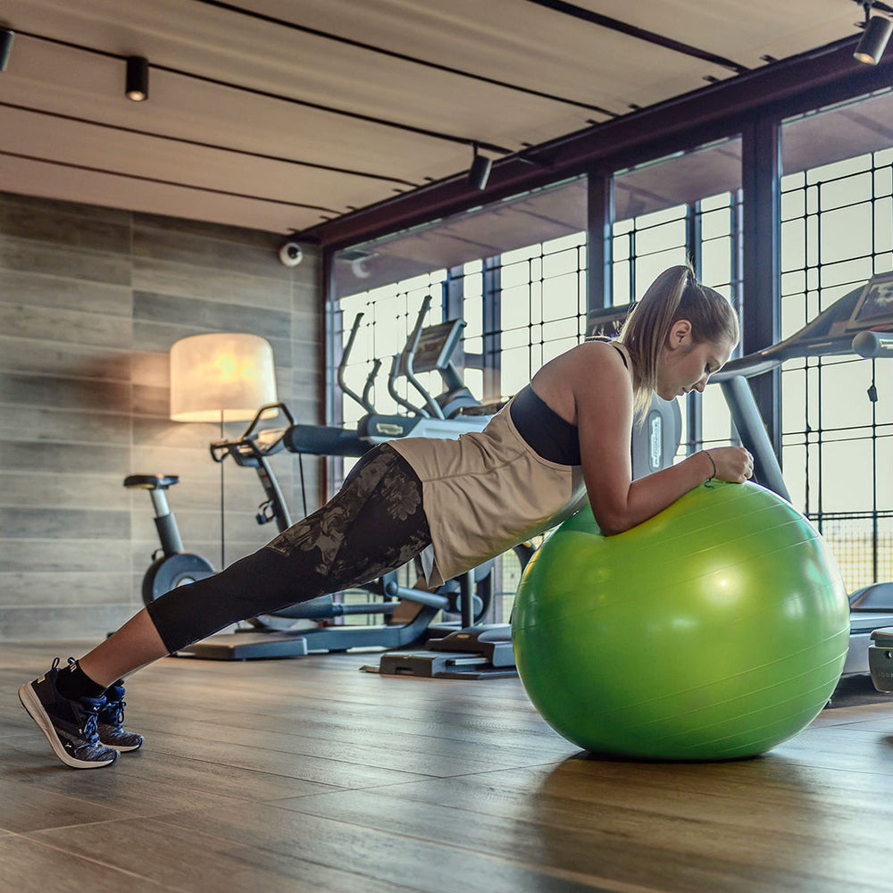Woman performing plank exercise with green X-DISCO Gymnic Physio Plus BRQ Physiotherapy Ball (85cm) in gym setting.