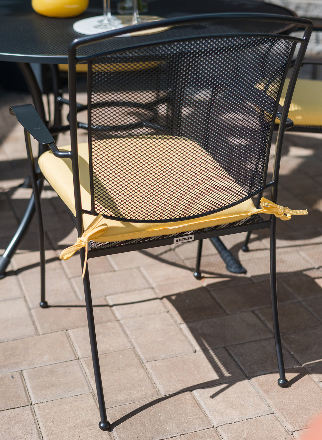 Henley Wrought Iron Mesh Stackable Arm Chair - Set Of 4 with iron grey mesh back and yellow cushion on patio.