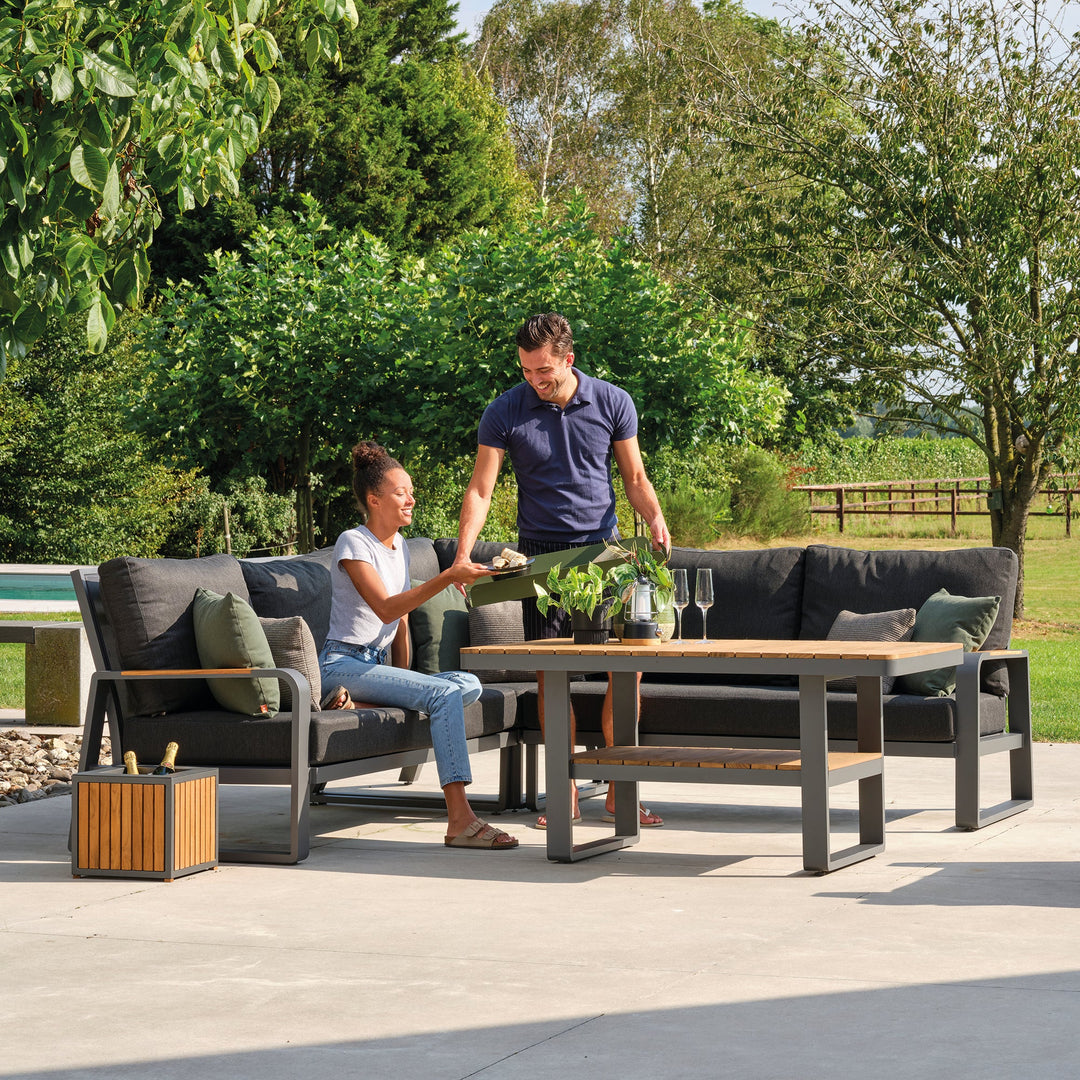 Montana Right Sofa with charcoal cushions and teak accents in a sunny outdoor garden setting with two people.