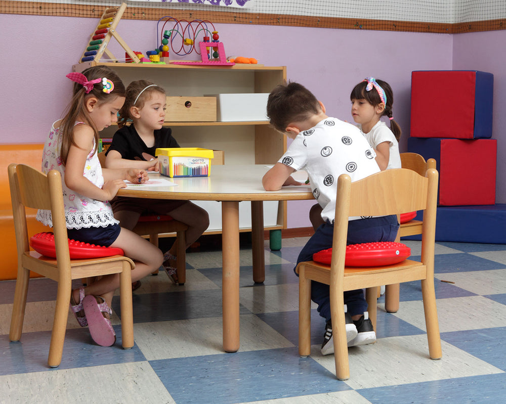 Children sitting on red Gymnic Disc 'O' Sit Jr. cushions at a small table in a classroom setting.