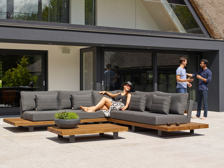 Fitz Roy Teak Corner Set with charcoal cushions and teak wood frame on patio with people relaxing and chatting nearby