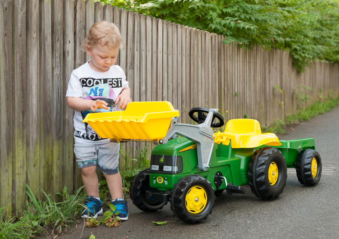 Toddler playing with John Deere Pedal Tractor With Loader featuring a yellow front loader and detachable trailer outdoors.