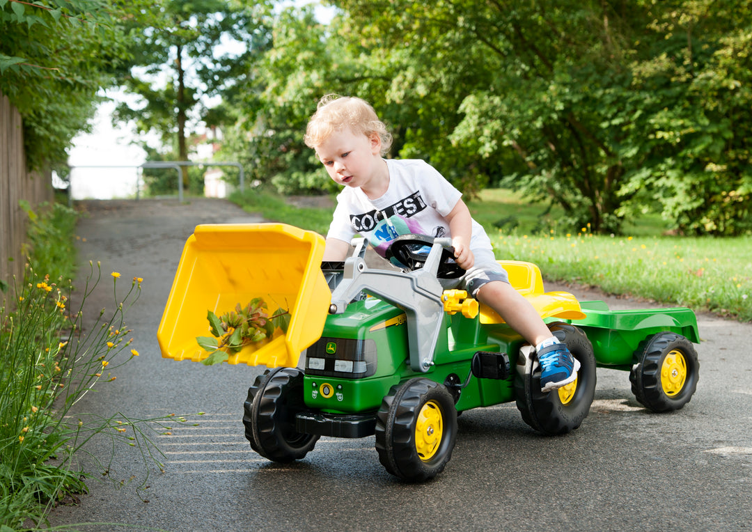 Child playing outdoors on a green John Deere Pedal Tractor With Loader with detachable trailer on a paved path.