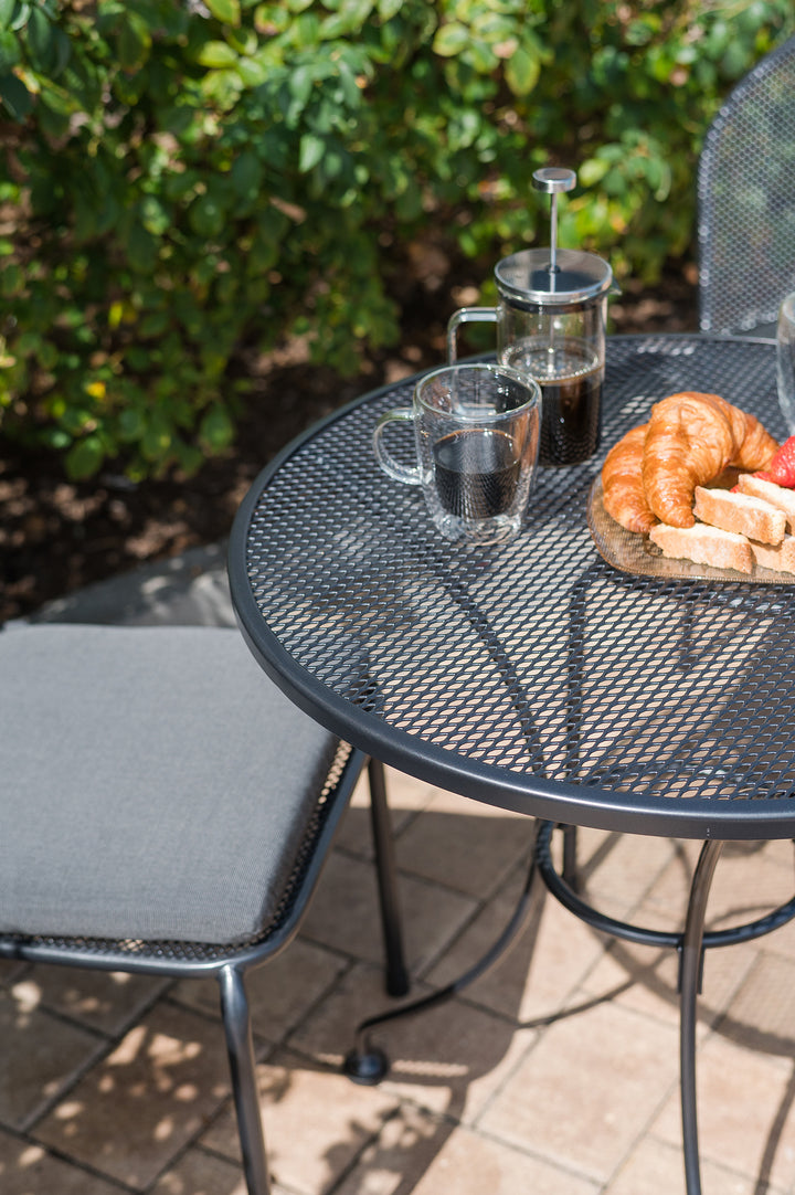 24-inch Round Mesh Top Table in iron grey with electrotherm coating, holding coffee and croissants outdoors.
