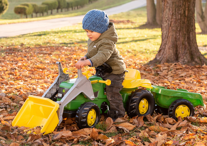 Child playing outdoors on a John Deere Pedal Tractor With Loader among autumn leaves near a tree.