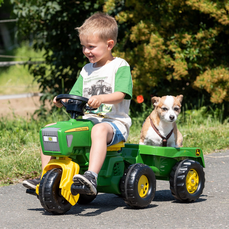 Child riding John Deere 3-Wheeled Pedal Tractor With Trailer carrying a small dog outside on pavement.