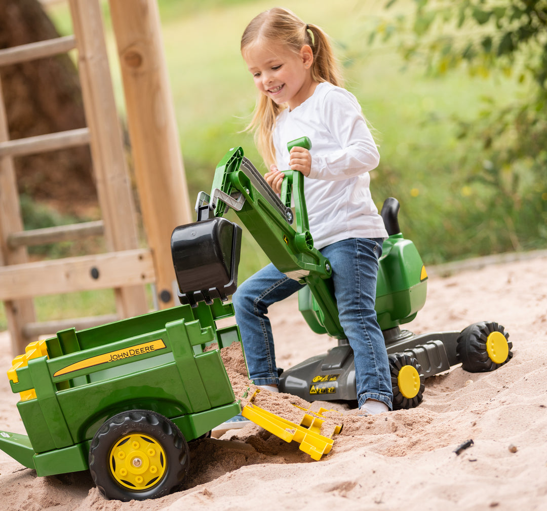 Child playing with the John Deere Ride-On Digger in the sand using hand-operated scoop and rotating arm.
