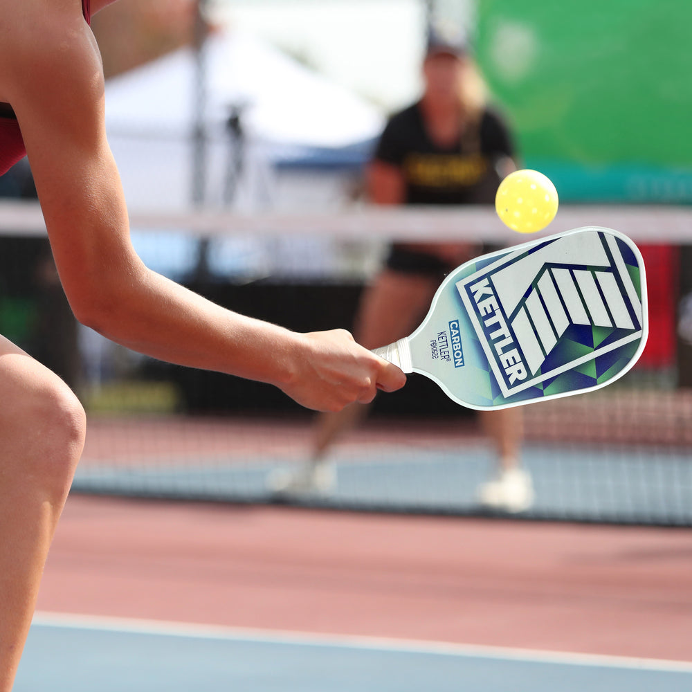 Player hitting a yellow ball with KETTLER® Carbon Tech Pickleball Set paddle during an outdoor game on court