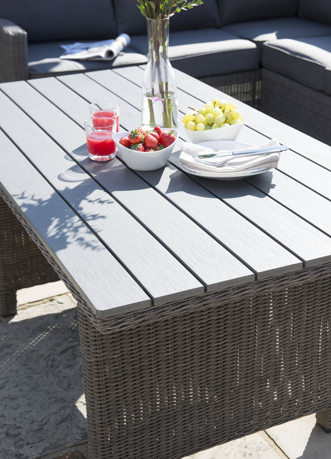 Palma Casual Dining Table with dark oak resin slats, handwoven rattan wicker base, and outdoor dining setup with fruit and drinks.