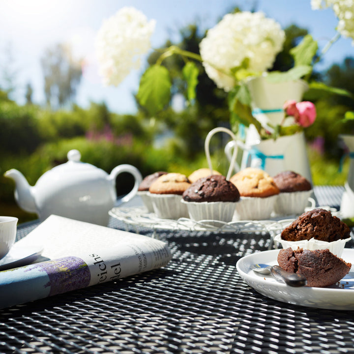60-inch Round Mesh Top Table with iron grey steel mesh surface holding muffins, teapot, and flowers outdoors